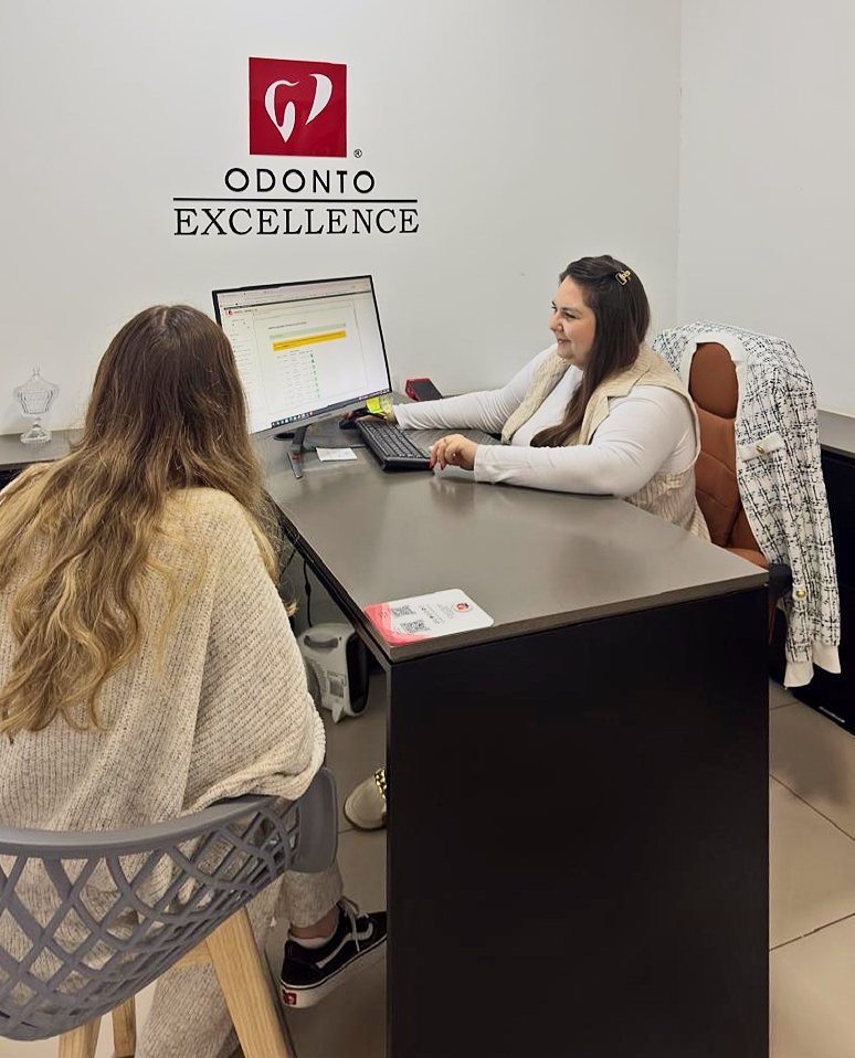Atendente da clínica conversando com uma paciente à mesa, com o logo da Odonto Excellence na parede